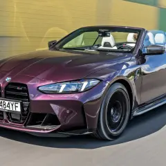 Fioletowy kabriolet BMW M4 F82 z białym wnętrzem pędzi po drodze, odbijając niebo i żółto-zieloną ścianę.