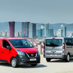Dwa samochody dostawcze Nissan NV300, czerwony i srebrny, stoją na tle panoramy miasta z nowoczesnymi budynkami.
