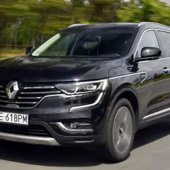 Czarny Renault Koleos 2.0 dCi pędzi po drodze, otoczony rozmytym zielonym krajobrazem.