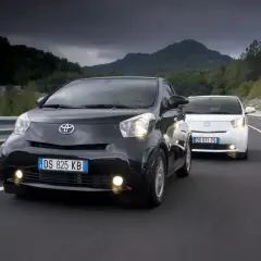 Dwa samochody Toyota iQ, czarny i biały, jadące po drodze w malowniczym, górzystym krajobrazie.