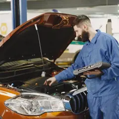 Mechanik w niebieskim kombinezonie sprawdza silnik pomarańczowego samochodu za pomocą urządzenia diagnostycznego.