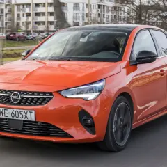 Pomarańczowy Opel Corsa 1.2 z czarnym dachem jedzie po drodze, w tle budynki mieszkalne.