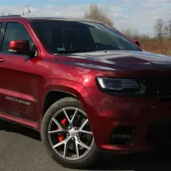 Czerwony Jeep Grand Cherokee SRT z czarnymi felgami i czerwonymi zaciskami hamulcowymi, zaparkowany na drodze wśród suchych traw i drzew.