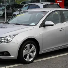 Srebrny Chevrolet Cruze 1.8 benzyna z napisem "HUPPERT" na masce, zaparkowany przed salonem samochodowym.