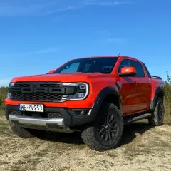 Pomarańczowy Ford Ranger Raptor gotowy na przygodę na piaszczystym terenie, z lasem w tle i błękitnym niebem.