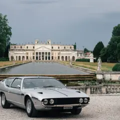 Srebrny Lamborghini Espada przed okazałą willą z basenem i rzeźbami. Klasyk motoryzacji w malowniczej scenerii.