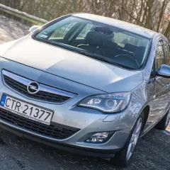 Srebrny Opel Astra J z przodu, zaparkowany na drodze z drzewami w tle.