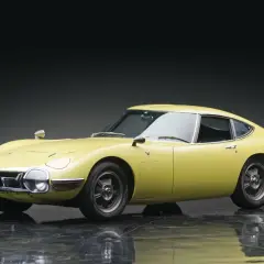 Żółta Toyota 2000GT z lat 60. prezentuje swoje klasyczne linie na błyszczącej podłodze.