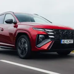 Czerwony Hyundai Tucson 1.6 T-GDI N Line pędzi po drodze, otoczony zielonymi polami i drzewami.