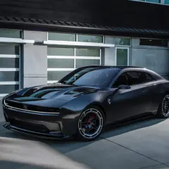 Nowoczesny, ciemnoszary Dodge Daytona z charakterystycznym pasem LED z przodu, zaparkowany przed nowoczesnym budynkiem z garażem.