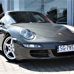 Szary Porsche 997 z czerwonym wnętrzem, zaparkowany na kostce brukowej przed budynkiem z lustrzaną fasadą.