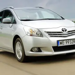 Srebrny Toyota Verso S jedzie po drodze, otoczony zielonymi polami.