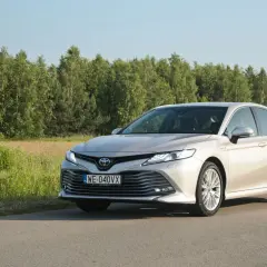 Elegancka Toyota Camry 2025 w kolorze szampańskim na tle zielonego krajobrazu.