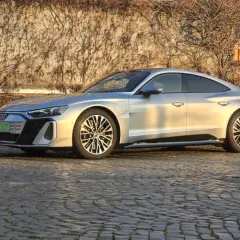 Srebrny Audi e-tron GT lśni w słońcu na brukowanej ulicy, z pnączami na ścianie w tle.