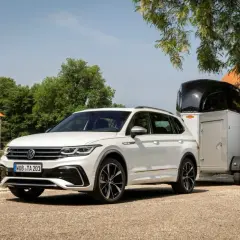 Biały Volkswagen Tiguan, jeden z wielu wszechstronnych volkswagen modele, holuje przyczepę z koniem przed zabytkowym budynkiem.