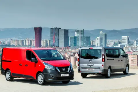 Dwa samochody dostawcze Nissan NV300, czerwony i srebrny, stoją na tle panoramy miasta z nowoczesnymi budynkami.