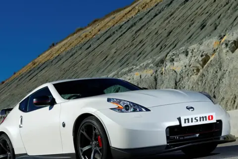 Biały Nissan 370Z Nismo z czerwonymi akcentami na lusterkach i zaciskach hamulcowych, zaparkowany na tle skalistego zbocza.