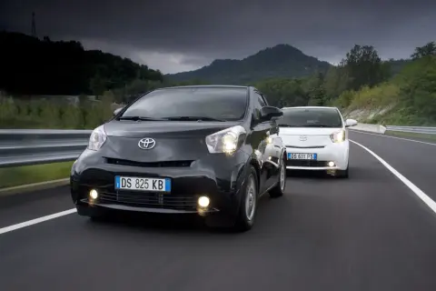 Dwa samochody Toyota iQ, czarny i biały, jadące po drodze w malowniczym, górzystym krajobrazie.