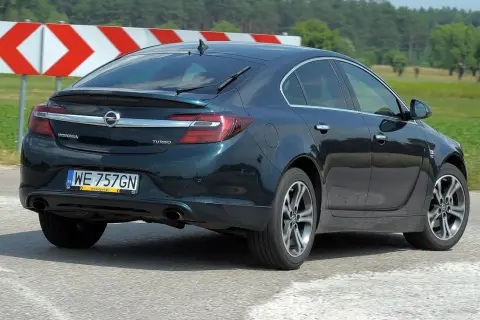 Ciemnozielony Opel Insignia 1.6 Turbo jedzie po drodze, za nim widać znaki ostrzegawcze i zielone pola.