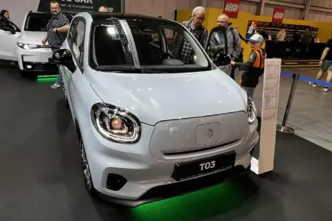 Biały, miejski samochód elektryczny MG T03 na wystawie. W tle widać innych zwiedzających i stoisko LEGO.