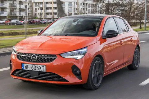 Pomarańczowy Opel Corsa 1.2 z czarnym dachem jedzie po drodze, w tle budynki mieszkalne.