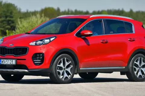 Czerwony Kia Sportage IV z kierowcą w okularach przeciwsłonecznych jedzie po drodze, otoczony zielenią.