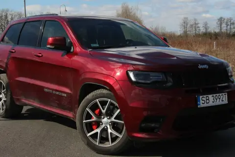 Czerwony Jeep Grand Cherokee SRT z czarnymi felgami i czerwonymi zaciskami hamulcowymi, zaparkowany na drodze wśród suchych traw i drzew.