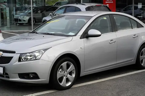 Srebrny Chevrolet Cruze 1.8 benzyna z napisem "HUPPERT" na masce, zaparkowany przed salonem samochodowym.