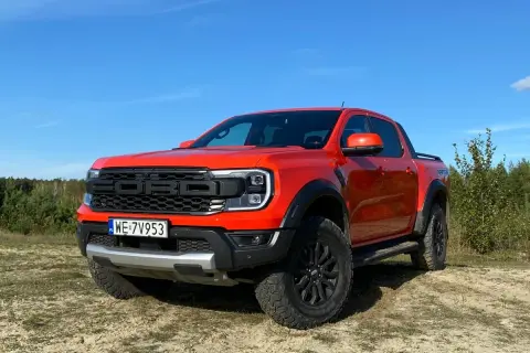 Pomarańczowy Ford Ranger Raptor gotowy na przygodę na piaszczystym terenie, z lasem w tle i błękitnym niebem.