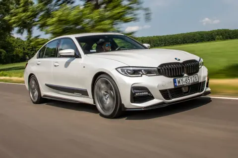 Białe BMW 320d xDrive pędzi po drodze, otoczone zielenią pól i drzew.