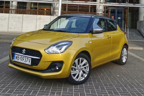 Złoty Suzuki Swift na tle nowoczesnego budynku. Wiele pozytywnych suzuki swift opinie potwierdza jego miejski charakter.
