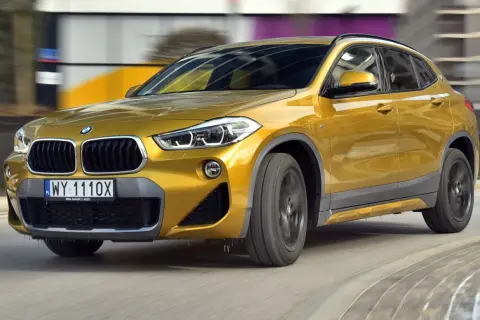 Złoty BMW F39 w ruchu, z widocznym przodem i bokiem, na tle rozmytego miejskiego krajobrazu.