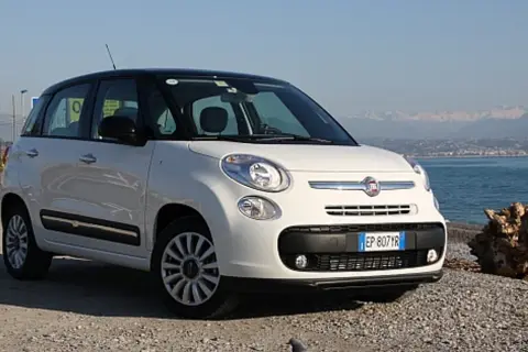 Biały fiat 500 l z czarnym dachem stoi na kamienistej plaży nad morzem.