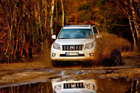 Biały Land Cruiser 100 szaleje po błotnistej drodze w lesie, wzbijając fontanny brudu.