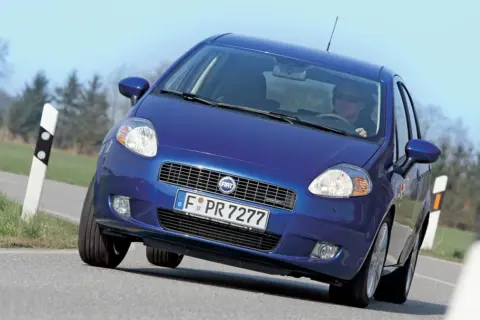 Niebieski Fiat Punto Grande pokonuje zakręt na drodze, z kierowcą w okularach za kierownicą.