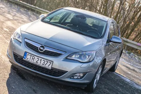 Srebrny Opel Astra J z przodu, zaparkowany na drodze z drzewami w tle.