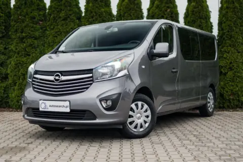 Szary Opel Vivaro, jeden z popularnych modeli Opla, zaparkowany przed żywopłotem.