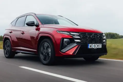 Czerwony Hyundai Tucson 1.6 T-GDI N Line pędzi po drodze, otoczony zielonymi polami i drzewami.