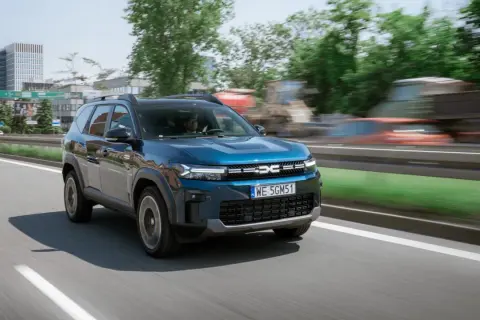 Niebieski Dacia Jogger na drodze, z widocznymi budynkami w tle. Marka samochodu na m.