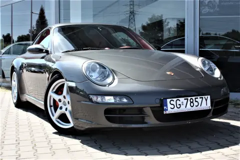 Szary Porsche 997 z czerwonym wnętrzem, zaparkowany na kostce brukowej przed budynkiem z lustrzaną fasadą.