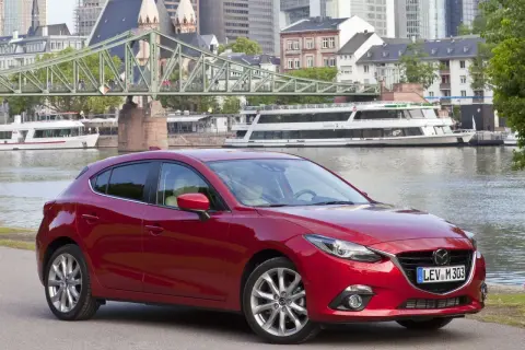 Czerwona Mazda 3 na tle miasta i rzeki. Wiele osób szuka takich aut, bo mazda 3 opinie ma zazwyczaj bardzo dobre.