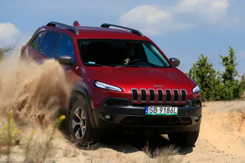 Czerwony Jeep Cherokee Trailhawk z impetem przemierza piaszczysty teren, wzbijając tumany piasku.