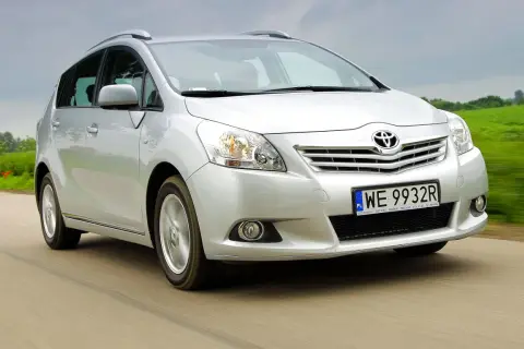 Srebrny Toyota Verso S jedzie po drodze, otoczony zielonymi polami.