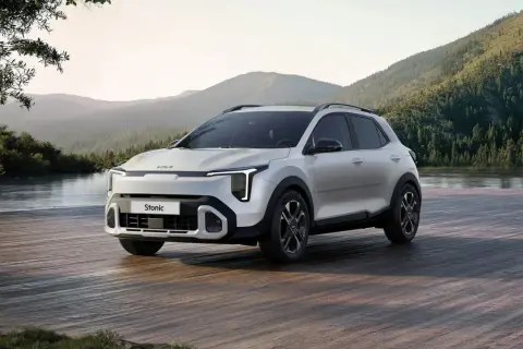 Nowy Kia Stonic 2024, biały SUV, zaparkowany na drewnianym pomoście nad jeziorem, w otoczeniu gór i lasów.