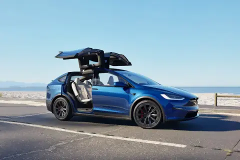 Niebieski Tesla Model X z otwartymi drzwiami typu falcon wing na tle plaży. Sprawdź aktualną tesla model x cena.