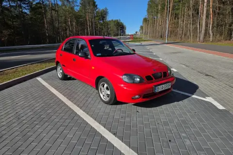 Czerwone Daewoo Lanos na parkingu z kostki brukowej, w tle las i błękitne niebo.
