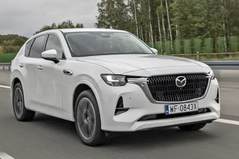 Biała Mazda CX-60 jedzie po drodze, otoczona drzewami. Sprawdź, jaka jest mazda cx 60 cena i wyposażenie.
