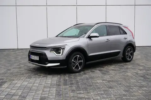 Srebrny Kia Niro na tle białej ściany. Wiele osób szuka kia niro opinie przed zakupem.