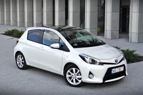 Biały Toyota Yaris z czarnym dachem, zaparkowany przed nowoczesnym budynkiem. Wiele osób szuka takich aut, czytając toyota yaris opinie.