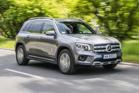 Szary Mercedes GLB 200 jedzie po drodze, otoczony zielenią drzew.